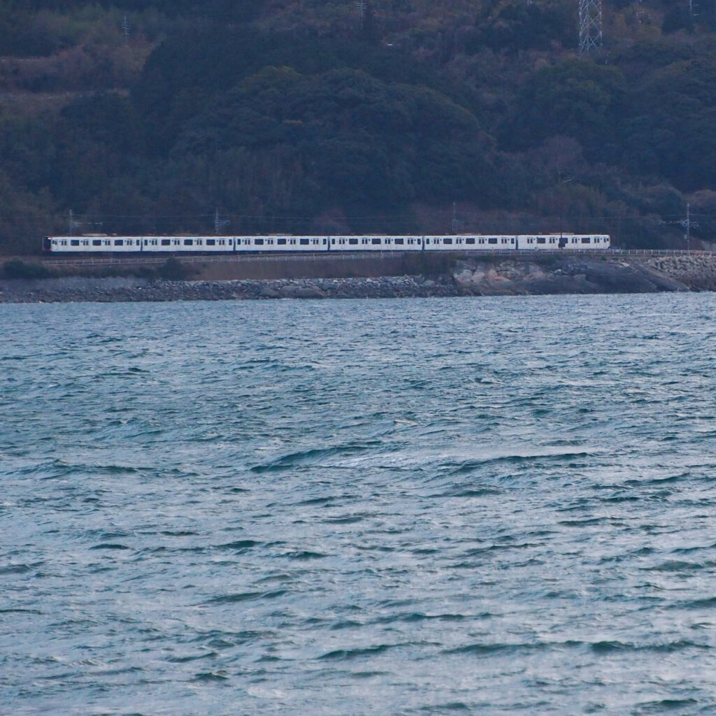 糸島市深江海岸近くから望遠で撮影した白い電車と青い海、山を背景に静かに走る列車の風景