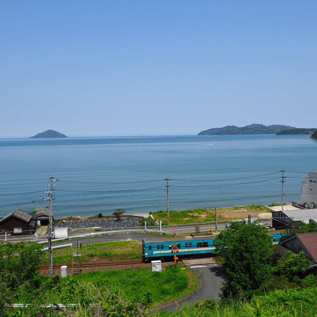 同じく二丈福井の高台から撮影、青い電車が踏切を渡る瞬間と青々とした海の広がり